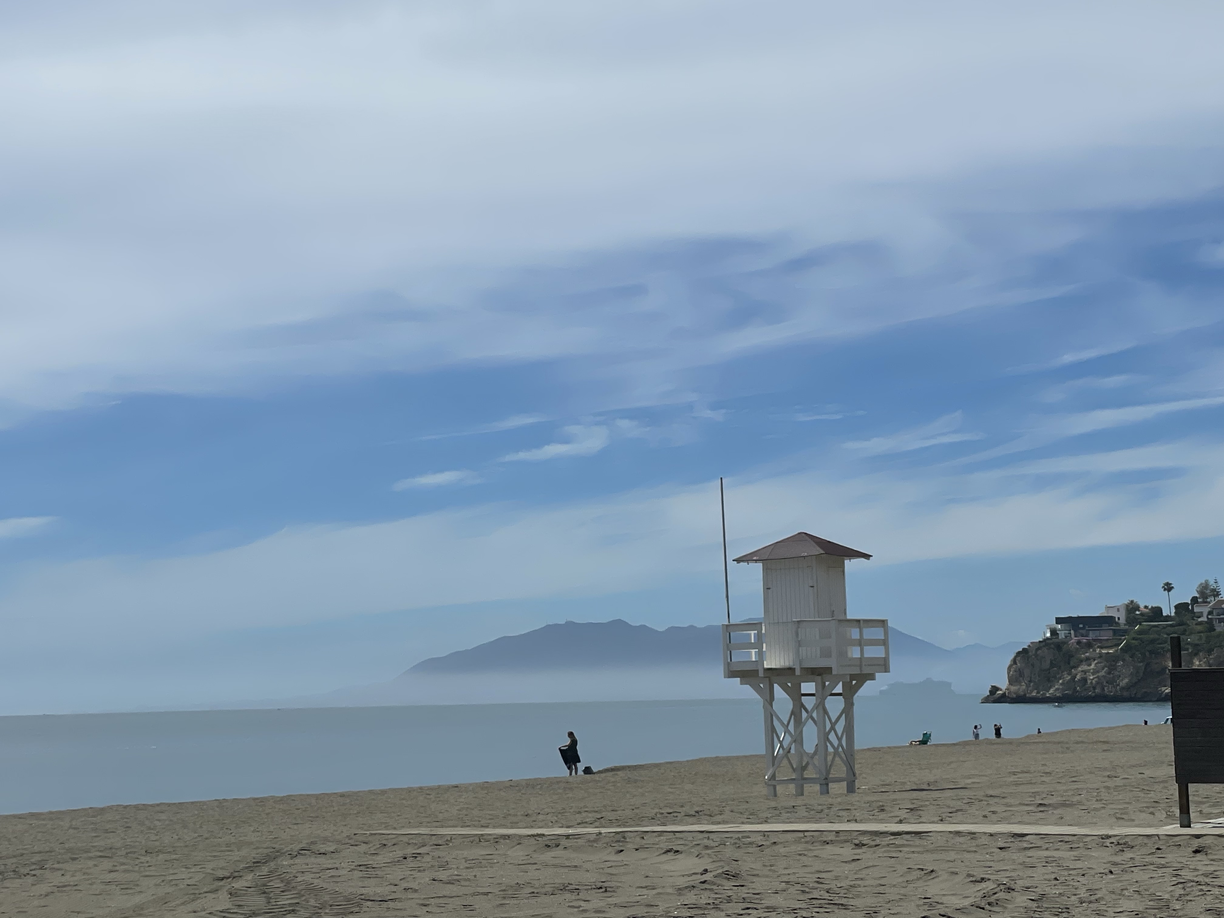 A serene beach scene featuring a lifeguard tower, a person walking with a dog, and distant mountains under a cloudy sky.